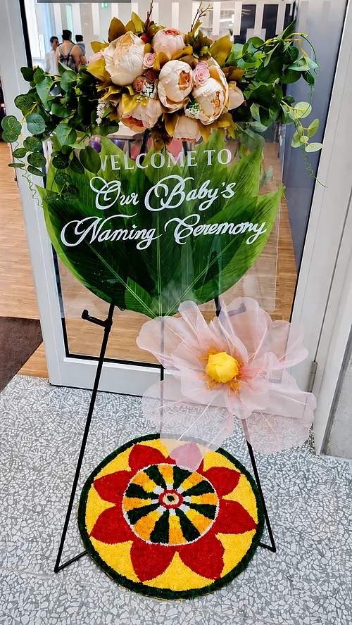 Baby naming ceremony welcome sign with floral arrangement, green leaf backdrop, decorative flower and colourful rangoli